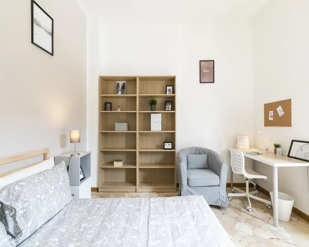 Camera da letto luminosa, arredata in stile soft con letto matrimoniale, scaffalatura in legno, poltrona accogliente e una piccola scrivania — decor pulito e neutro perfetto per annunci.