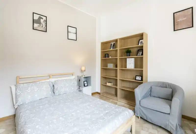 Camera da letto luminosa e semplice con letto matrimoniale, lampada da comodino, libreria in legno e una poltrona comoda — arredamento neutro e pulito, perfetto per un annuncio.