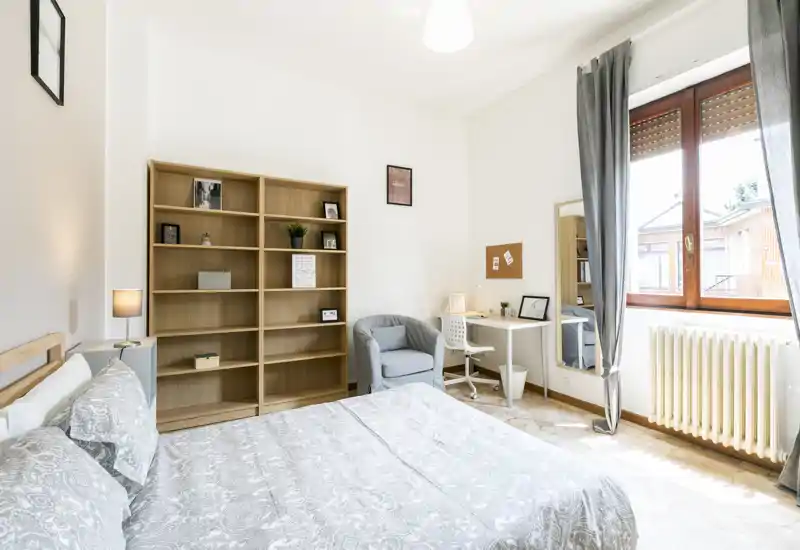 Camera da letto luminosa e ben arredata con letto matrimoniale in primo piano, libreria in legno, poltrona accogliente e una scrivania vicino a una grande finestra che restituisce molta luce naturale.