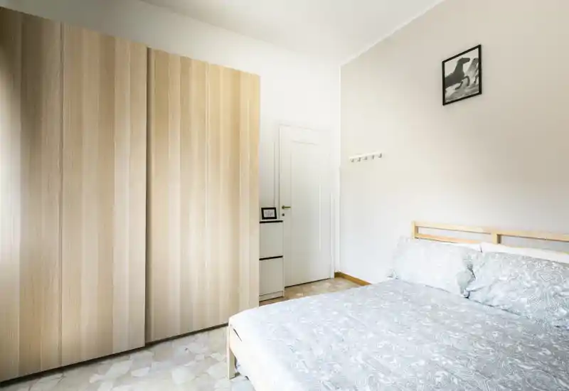 Camera da letto luminosa e minimal con letto matrimoniale con biancheria dal motivo e un grande armadio in legno chiaro. Linee pulite, toni neutri e luce naturale creano un'atmosfera calma e accogliente, perfetta per annunci di affitto.