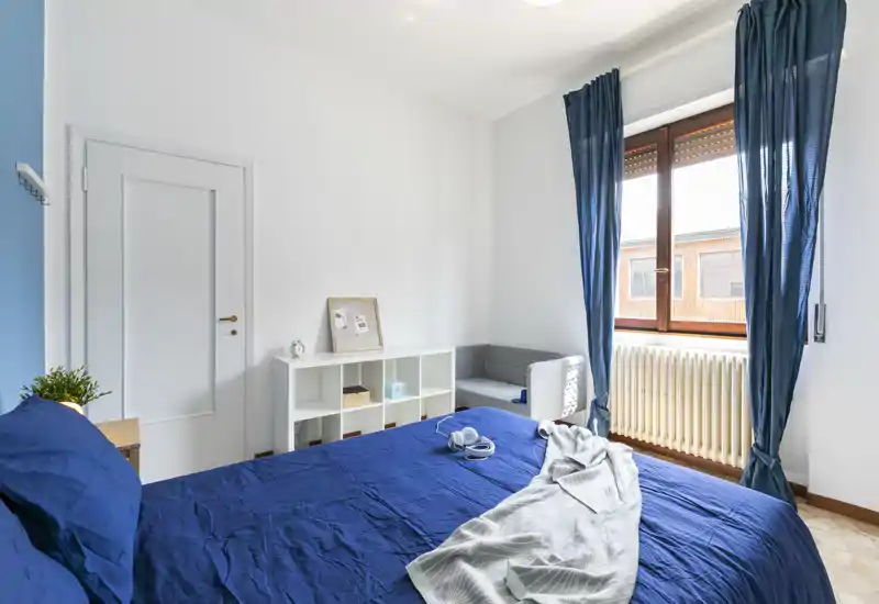 Bright, modern bedroom with a blue-dressed double bed, natural light from a large window and a small seating area. Clean, airy styling ideal for rental photos.