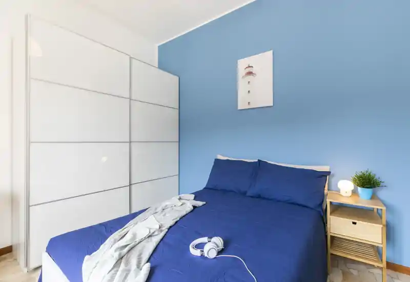 Bright, well-staged bedroom with a double bed dressed in deep blue linens, a small wooden nightstand with lamp and plant, and a white sliding-door wardrobe against a soft blue wall.
