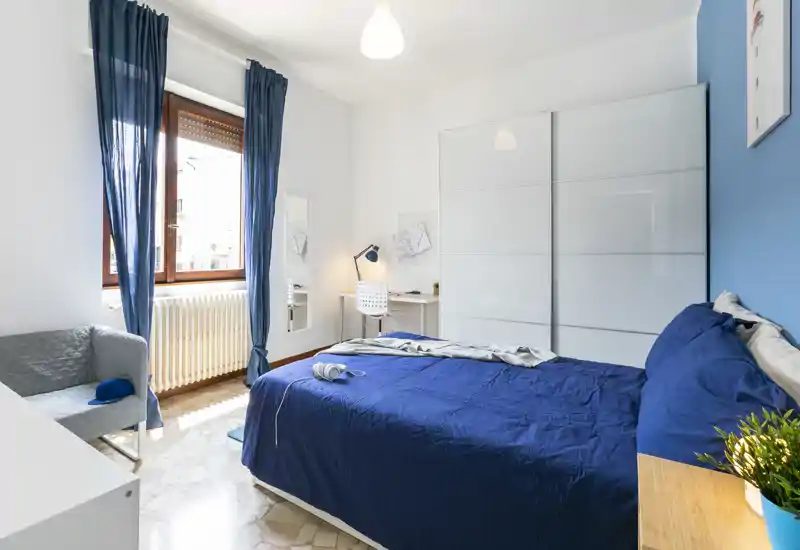 Bright, modern bedroom with a double bed dressed in deep blue linens, a large white wardrobe, a small desk and a comfortable chair by the window letting in natural light.