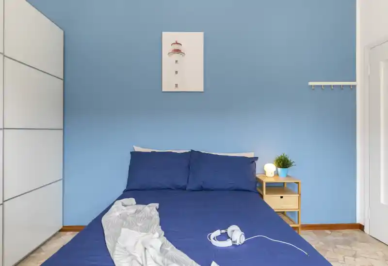 Bright, neatly styled bedroom with a double bed dressed in deep blue linens, a small wooden bedside table with lamp and plant, and a white wardrobe—calm coastal-inspired decor.