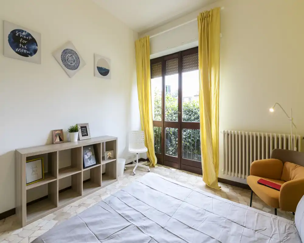 Chambre lumineuse avec lit double, rideaux jaunes et un fauteuil chaleureux près de la fenêtre. Étagères simples et décor mural apportent une ambiance accueillante, parfaite pour une annonce locative.