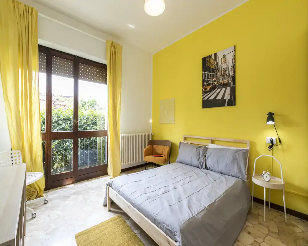 Chambre lumineuse et moderne avec un lit double, linge de lit gris et un mur d'accent jaune vif. Grande baie vitrée apportant beaucoup de lumière naturelle et un fauteuil pour un coin lecture confortable.
