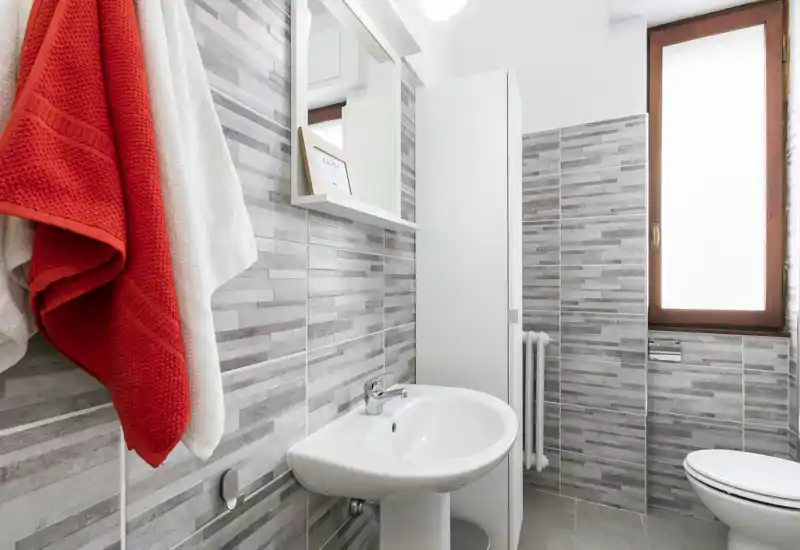 Bright, modern bathroom with a white pedestal sink, wall-mounted mirror cabinet, tiled walls and a window providing natural light.