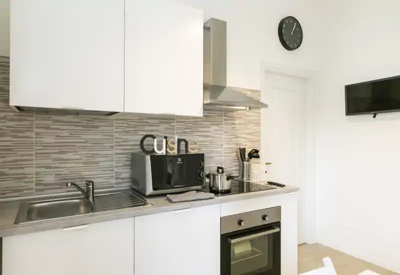 Compact modern kitchenette with stainless sink, induction hob and built-in oven. Neutral tiles and white cabinets create a clean, functional cooking area.