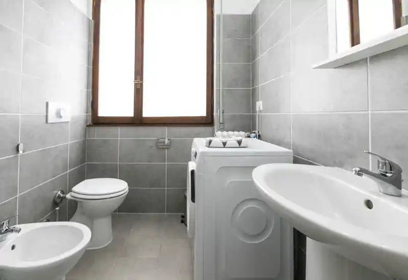 Clean modern bathroom with toilet, bidet, sink and a washing machine under a bright window; light gray tiles and chrome fixtures give a fresh, functional look.