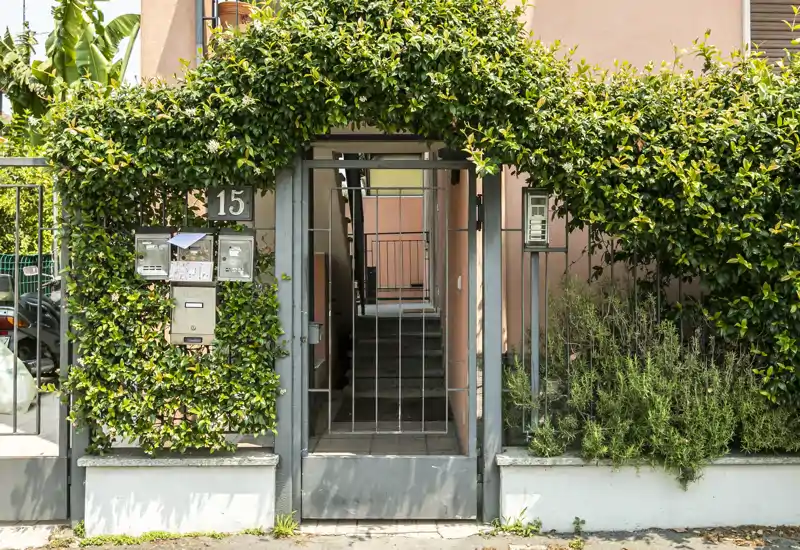 Sunny gated entrance framed by lush greenery, showing mailboxes, house number and an inner stairway — ideal to illustrate the building entry point.