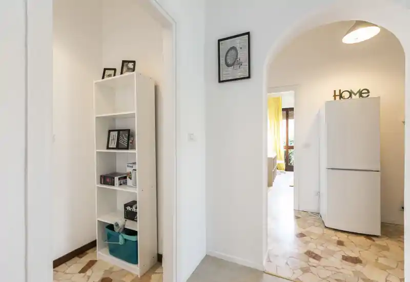 Bright, clean entrance hallway with decorative archways and a tall storage shelf; view into the kitchen area with a refrigerator and natural light from the room beyond.
