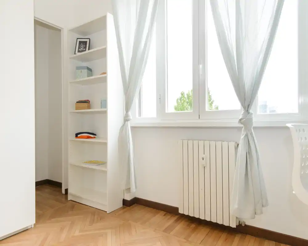 Angolo living luminoso e minimale con pavimento in parquet a spina di pesce, termosifone sotto una grande finestra e tende chiare; una libreria incassata offre spazio contenitivo.