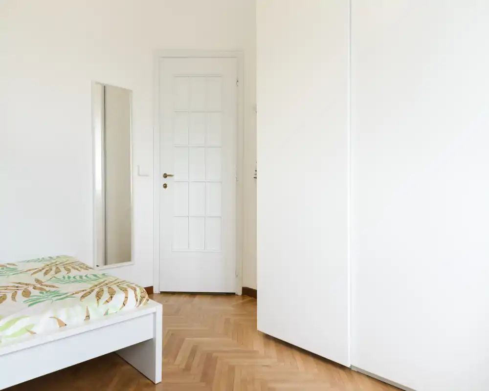 Camera da letto minimal e luminosa con struttura letto bianca, copriletto a fantasia e pavimento in parquet naturale; stile pulito e moderno con buona luce naturale.