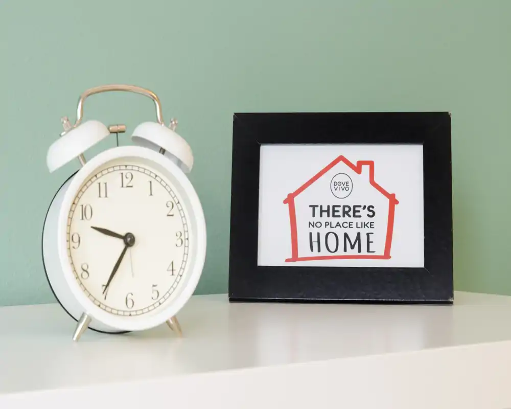 Primo piano di una sveglia decorativa e una cornice con la scritta "There's no place like home" su una superficie chiara contro una parete verde menta. La foto mette in risalto gli accessori e lo styling, non una stanza specifica.