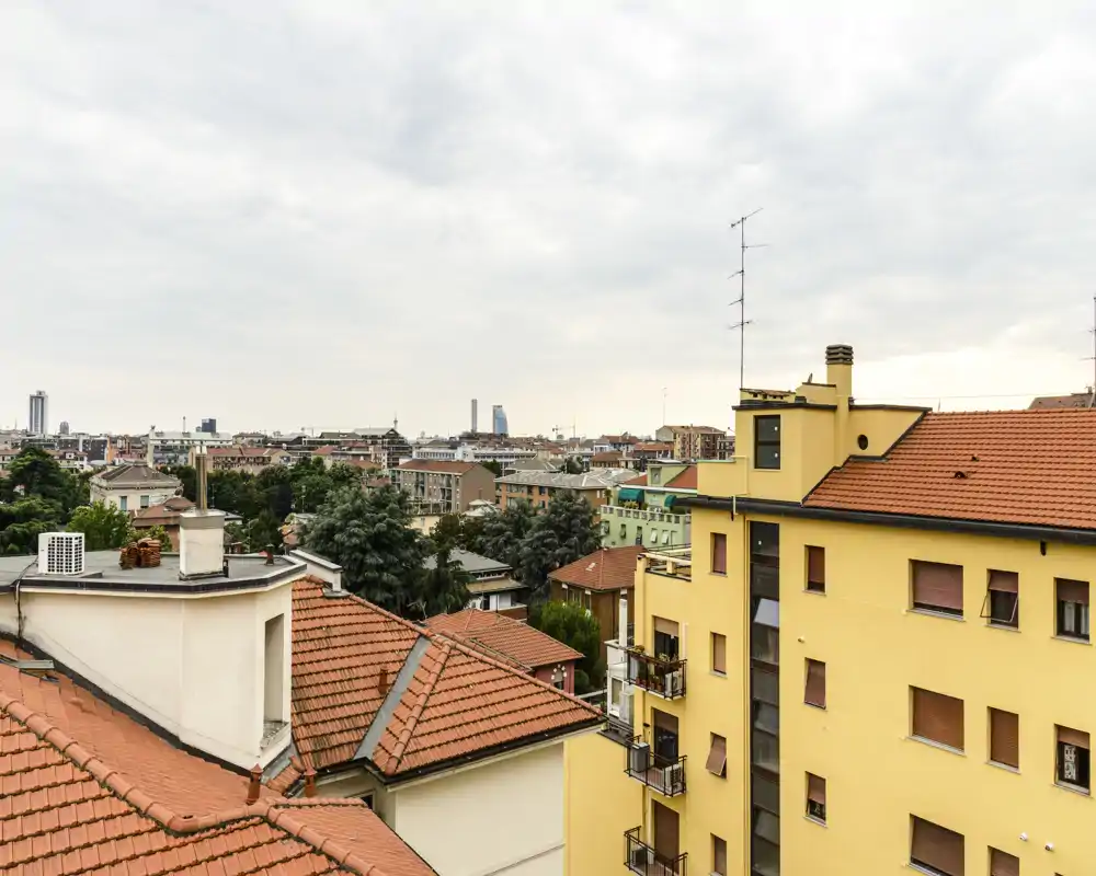 Ampia vista dai tetti su un quartiere residenziale con coperture in cotto e lo skyline cittadino in lontananza sotto una luce soffusa e nuvolosa.