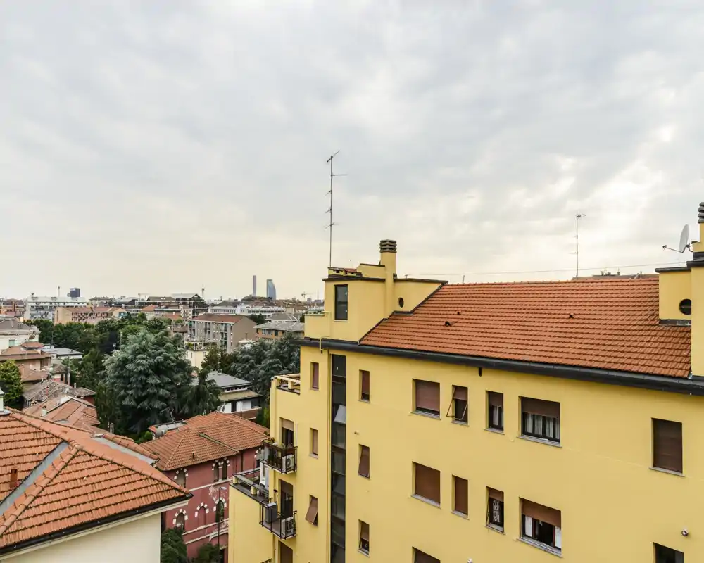 Vista sui tetti e sugli edifici circostanti da un balcone/terrazza; tetti in tegole calde e skyline lontano sotto un cielo leggermente nuvoloso.