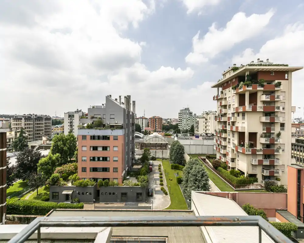 Vista cittadina da un balcone con palazzi residenziali, cortili verdi e cielo parzialmente nuvoloso — perfetto per mettere in evidenza lo spazio esterno e la vista sul quartiere.