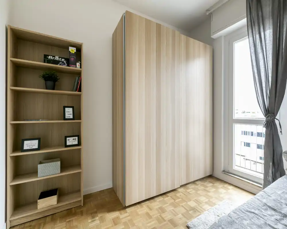 Angolo di camera da letto luminoso con un grande armadio in legno chiaro, scaffalatura aperta e una parziale vista del letto vicino alla finestra che lascia entrare luce naturale.