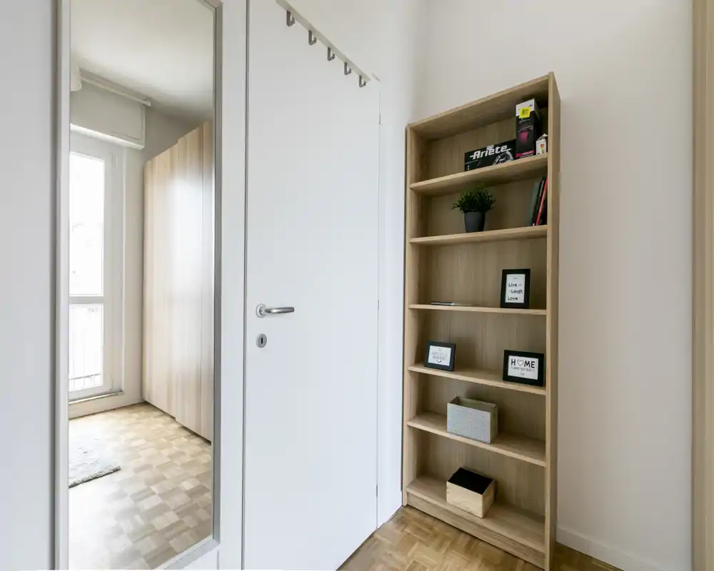 Ingresso luminoso e minimal con specchio a figura intera, porta bianca e una libreria in legno per riporre oggetti e decorazioni.