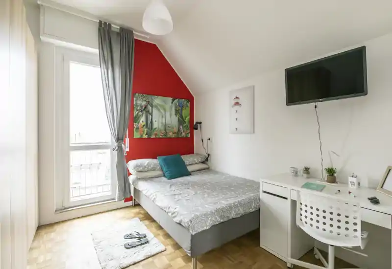 Camera da letto luminosa e ben arredata con letto matrimoniale, cuscini decorativi, quadri e una scrivania accanto alla TV a parete. Ampia finestra con luce naturale e pavimento in parquet che dona calore.