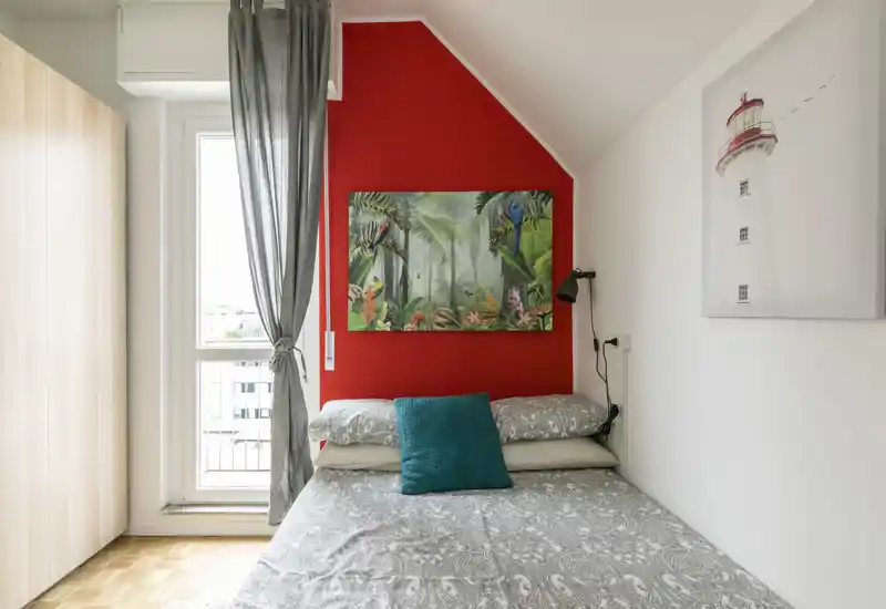 Camera da letto luminosa e compatta con letto matrimoniale contro una parete rossa, quadro tropicale decorativo e cuscino turchese. La luce naturale dalla finestra e l'armadio semplice rendono la stanza pratica e accogliente.