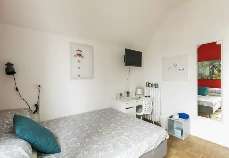 Camera da letto luminosa e ben allestita con un grande letto in primo piano, una scrivania compatta e TV a parete. Tessuti morbidi e arredi coordinati creano un'atmosfera accogliente, ideale per annunci.