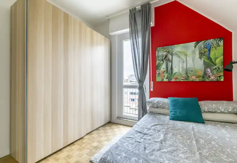 Camera da letto luminosa e moderna con grande armadio, biancheria a fantasia e parete d'accento colorata; luce naturale dalla finestra e quadro elegante rendono l'ambiente accogliente.