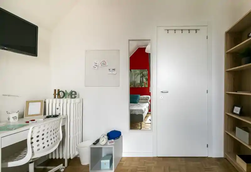 Camera da letto luminosa e ordinata visibile attraverso un'apertura: si intravede il letto con cuscini e copriletto decorato; in primo piano una scrivania compatta e scaffali.