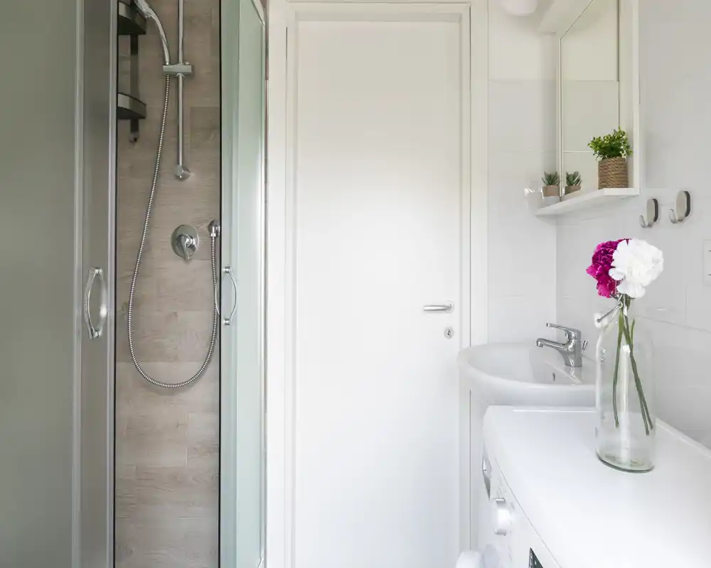Bagno compatto e luminoso con doccia con ante in vetro, lavabo bianco e piano della lavatrice con un vaso di fiori; finiture moderne e pulite.