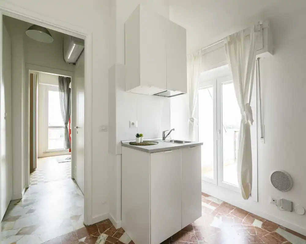 Angolo cottura compatto e moderno con mobile bianco, piccolo lavello e cappa vicino alla porta del balcone luminosa; stile minimal pulito e luce naturale danno una sensazione ariosa.