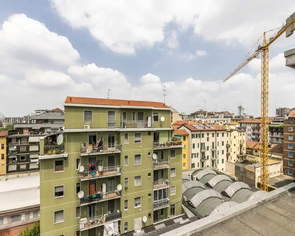 Vista cittadina da una terrazza o finestra con condomini vicini, tetti e una gru di cantiere sotto un cielo parzialmente nuvoloso.