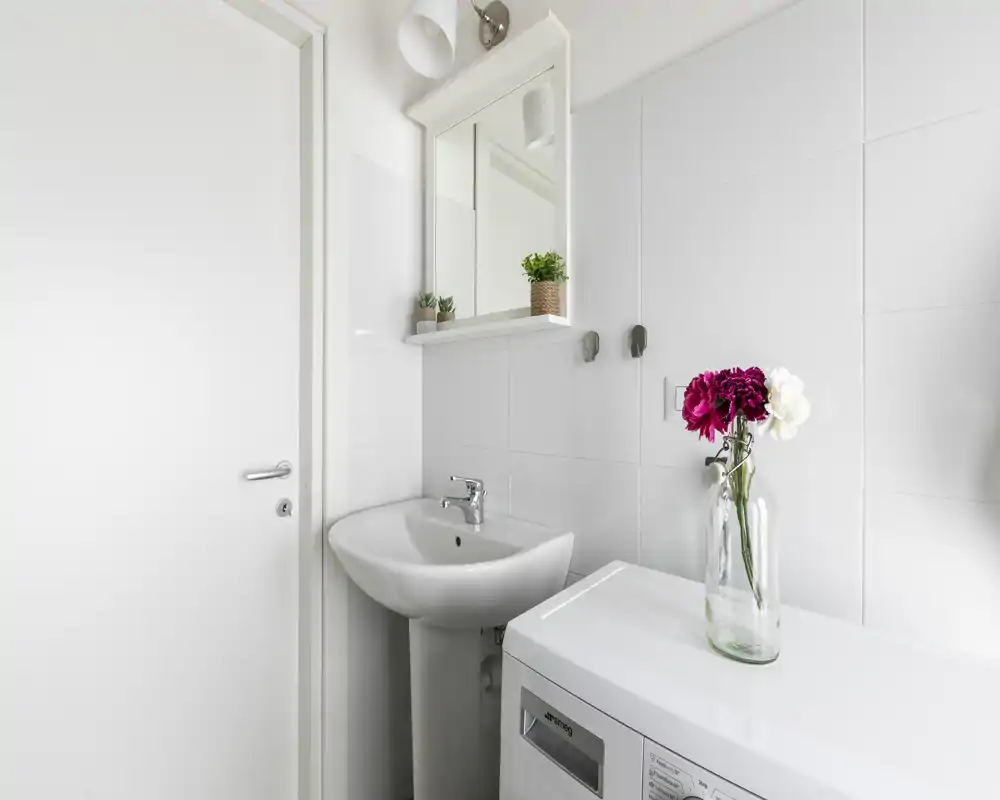 Angolo bagno moderno e luminoso con lavabo a colonna, specchiera con mobiletto e una lavatrice su cui è posizionato un vaso di vetro con fiori freschi.