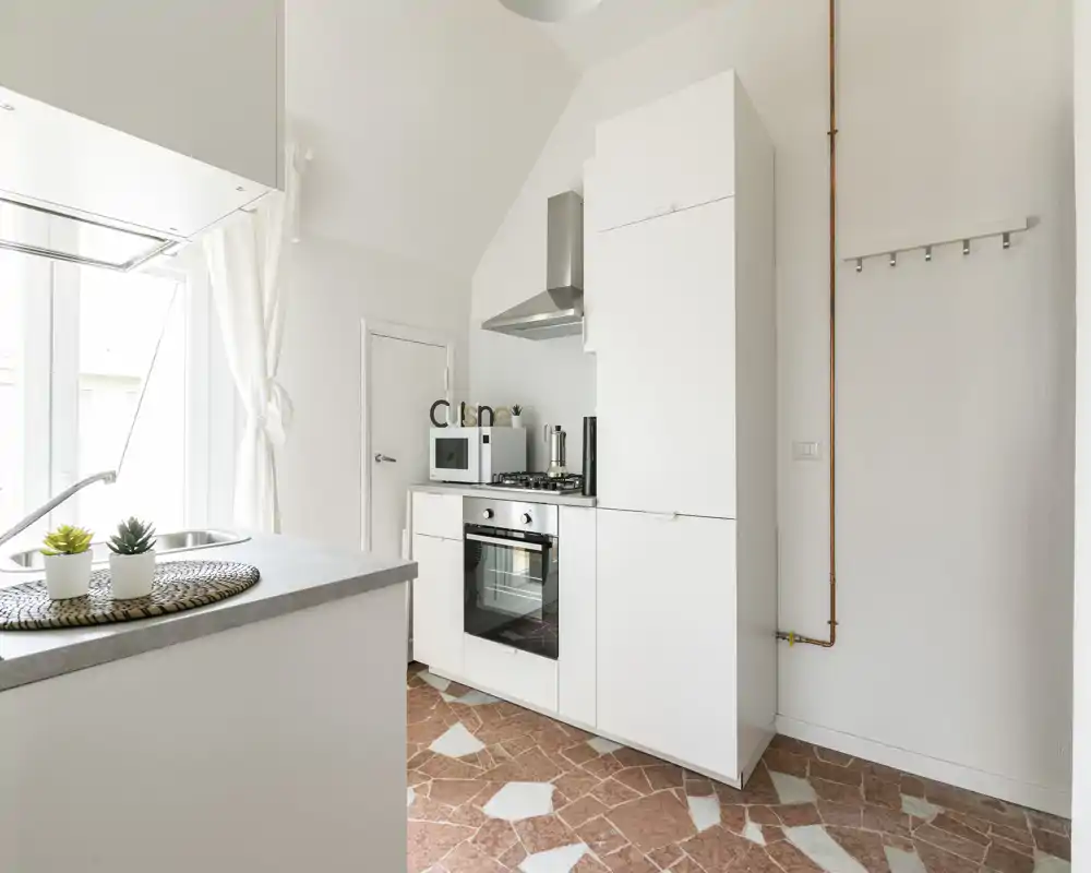 Cucina compatta e moderna con mobili bianchi, forno integrato e piano cottura, piccolo bancone con lavello — stile minimal pulito e luce naturale da una finestra ampia.