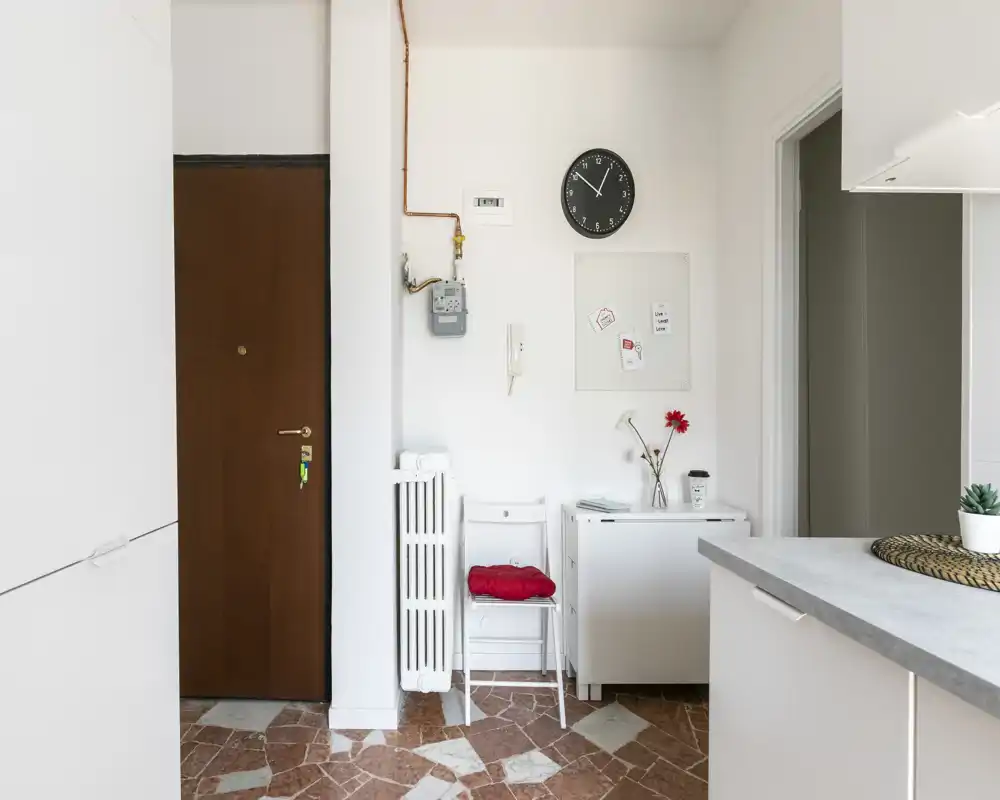 Ingresso appartamento piccolo e luminoso con porta d'ingresso in legno, termosifone e un mobiletto laterale compatto; decorazione neutra e pavimento in piastrelle per un ingresso pulito e accogliente.
