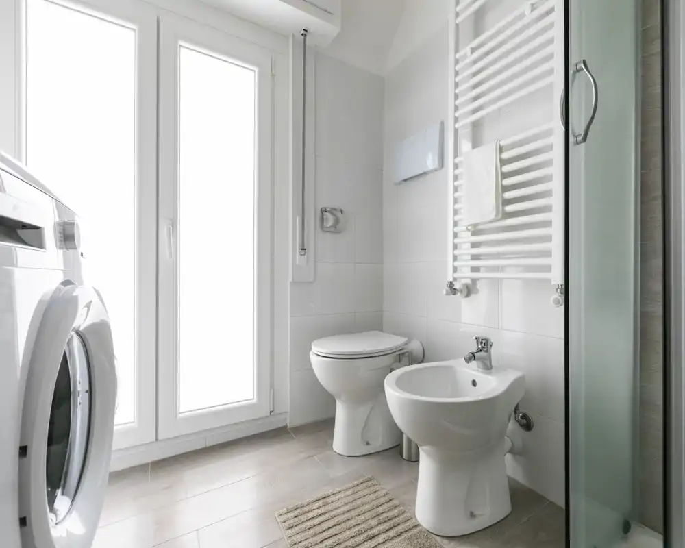 Bagno moderno e pulito con wc, bidet, box doccia in vetro e una lavatrice accanto a ampie finestre opache.