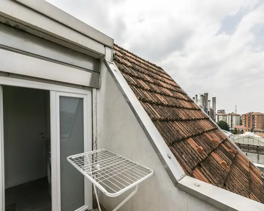 Piccolo balcone sul tetto con vista sulle tegole, porta scorrevole in vetro e stendibiancheria a parete; sullo sfondo si vede lo skyline urbano.