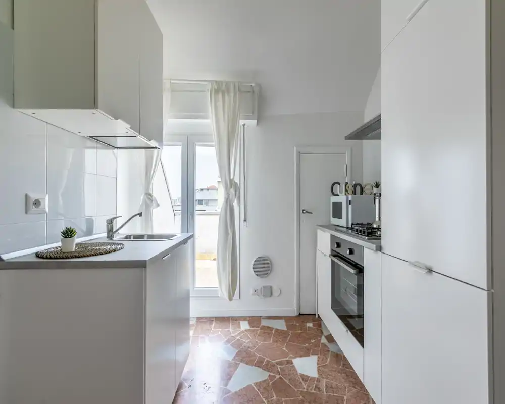 Cucina luminosa e compatta con mobili bianchi, forno incassato e lavello vicino a una porta finestra che affaccia all'esterno. Stile minimal e pulito, luce naturale e pavimento in piastrelle dal motivo terracotta.