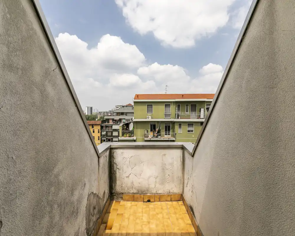 Piccolo balcone privato con pavimento piastrellato e alti muri laterali, con vista sugli edifici vicini e sul cielo — adatto come spazio esterno compatto o per deposito.