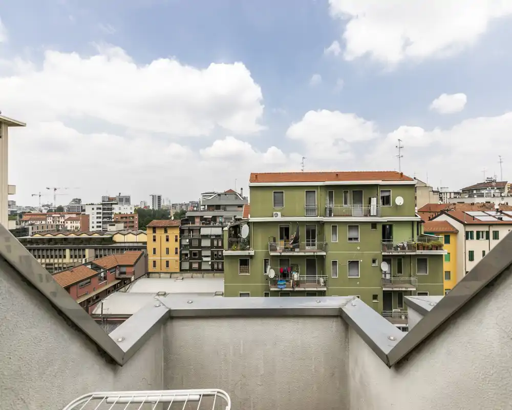 Piccolo balcone soleggiato con vista sulla città: parapetto rialzato e stendibiancheria in metallo in primo piano, con vista sui palazzi vicini e il cielo nuvoloso.