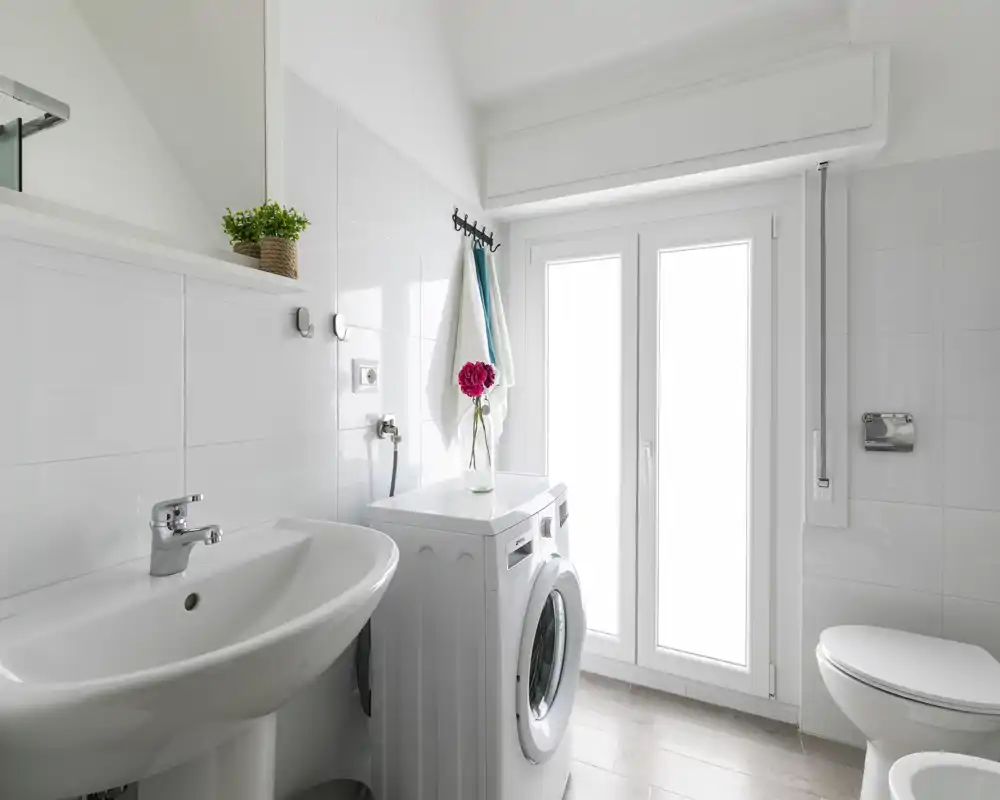 Bagno moderno e luminoso con lavabo, WC e una lavatrice a carica frontale accanto a una finestra satinata; piastrelle bianche e arredo minimale creano uno spazio funzionale e pulito.
