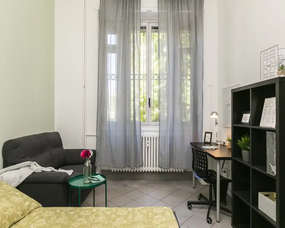 Camera da letto luminosa e ordinata con letto in primo piano, poltrona comoda, scrivania e scaffalatura — ideale per un single o affitti brevi.