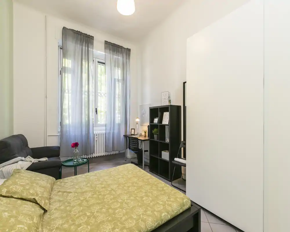 Camera da letto luminosa e ben arredata con un grande letto in primo piano, un divano comodo e una piccola postazione di lavoro vicino alla finestra; luce naturale morbida filtrata da tende leggere.