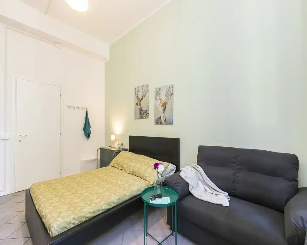 Camera da letto luminosa e ben allestita con letto matrimoniale, lampada da comodino, quadri alle pareti e divano comodo — perfetta per un monolocale accogliente.