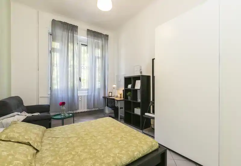 Dormitorio luminoso y bien decorado con una gran cama en primer plano, un sofá cómodo y una pequeña zona de escritorio junto a la ventana; luz natural suave a través de cortinas translúcidas.