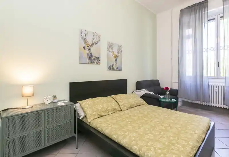 Dormitorio luminoso y acogedor con cama doble, mesita de noche y un sillón junto a la ventana. Paleta neutra, cuadros decorativos y textiles suaves que crean un ambiente agradable, perfecto para alquileres a corto plazo.