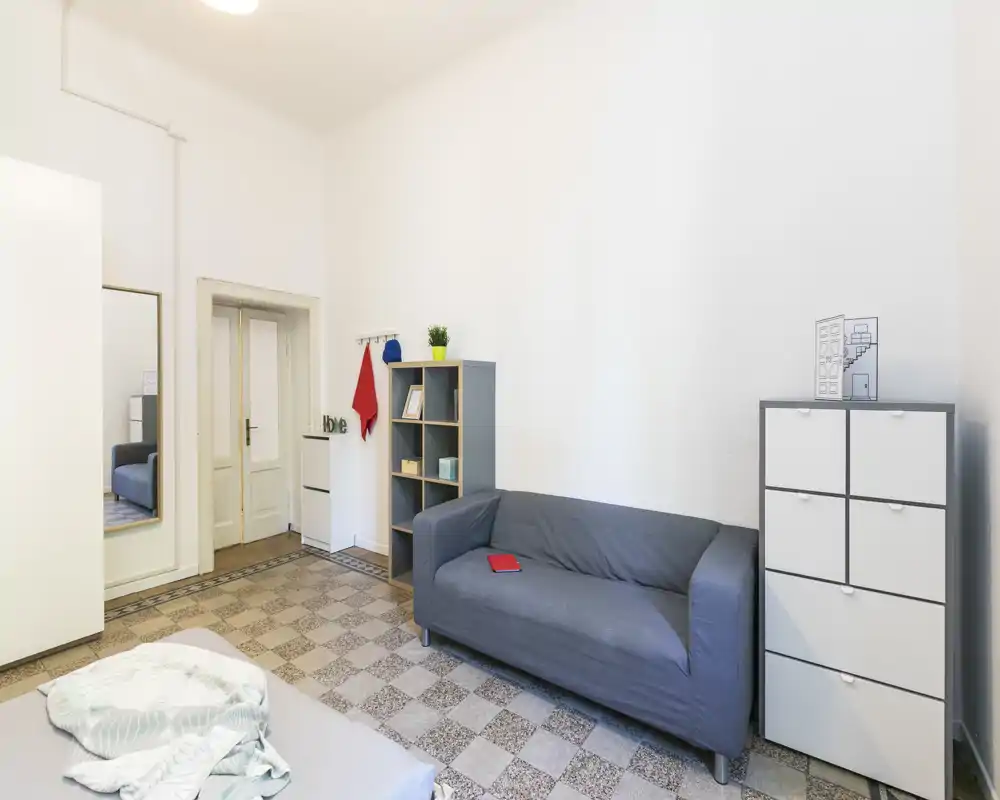 Dormitorio sencillo y luminoso con colchón visible en primer plano y una zona de estar con sofá, estantería y armarios.