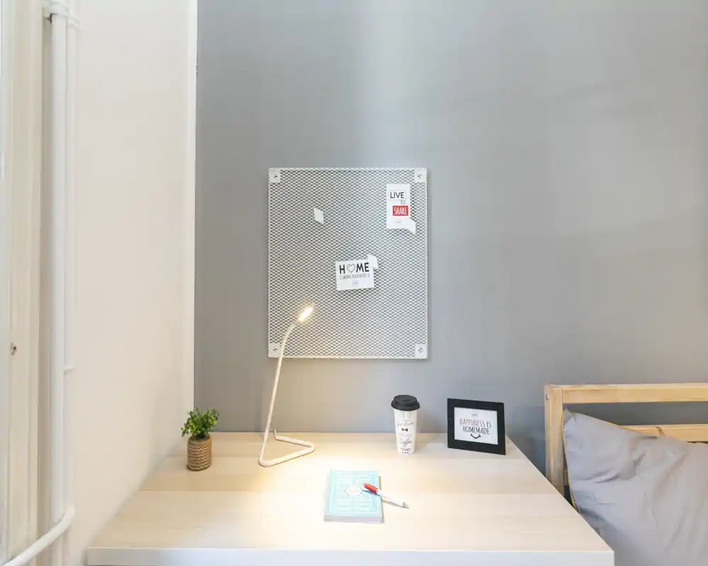 Rincón de dormitorio compacto con la estructura de la cama visible en el borde derecho y un escritorio ordenado con lámpara moderna, pequeña planta y tablero de notas encima.