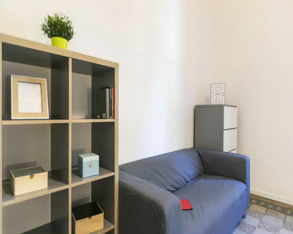Zona de estar compacta con un sofá gris de dos plazas, estantería con cajas decorativas y una planta — espacio luminoso y de estilo sencillo, ideal para relajarse.