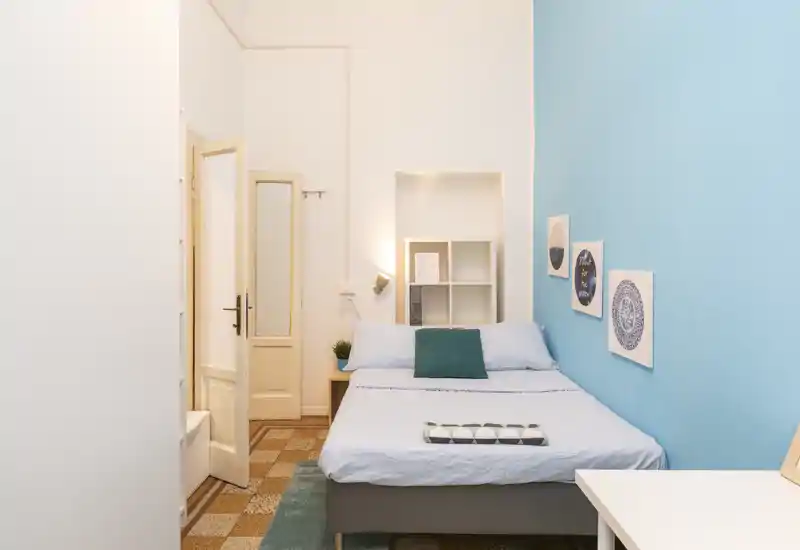 Dormitorio luminoso y compacto con una cama doble bien hecha frente a una pared azul, cuadros decorativos, estantería junto a la mesita y un pequeño escritorio — ideal para una estancia en la ciudad.