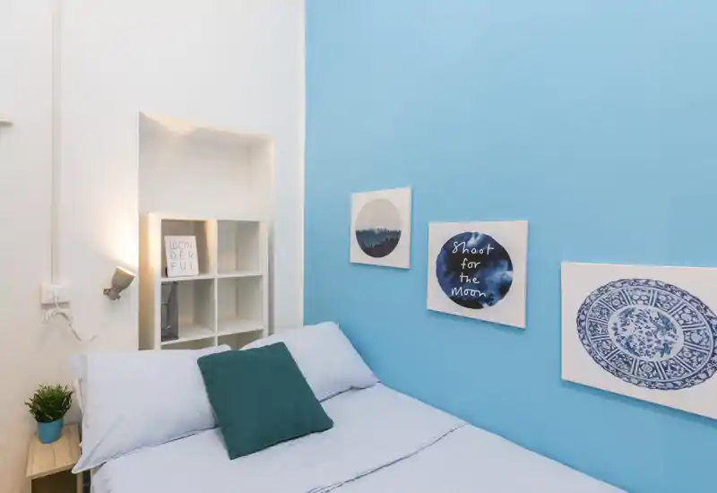 Dormitorio luminoso y moderno con pared azul claro, cama bien hecha con cojín verde, cuadros en la pared y pequeña estantería con luz tenue.